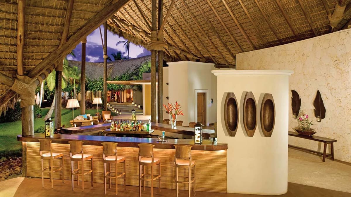 Canoa lobby bar with palapa roof, carved stone accents, and premium spirits under evening lighting