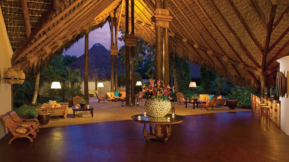 Open-air lobby with soaring palapa cathedral ceiling, tropical furniture, candlelit tables, and Canoa lobby bar at dusk