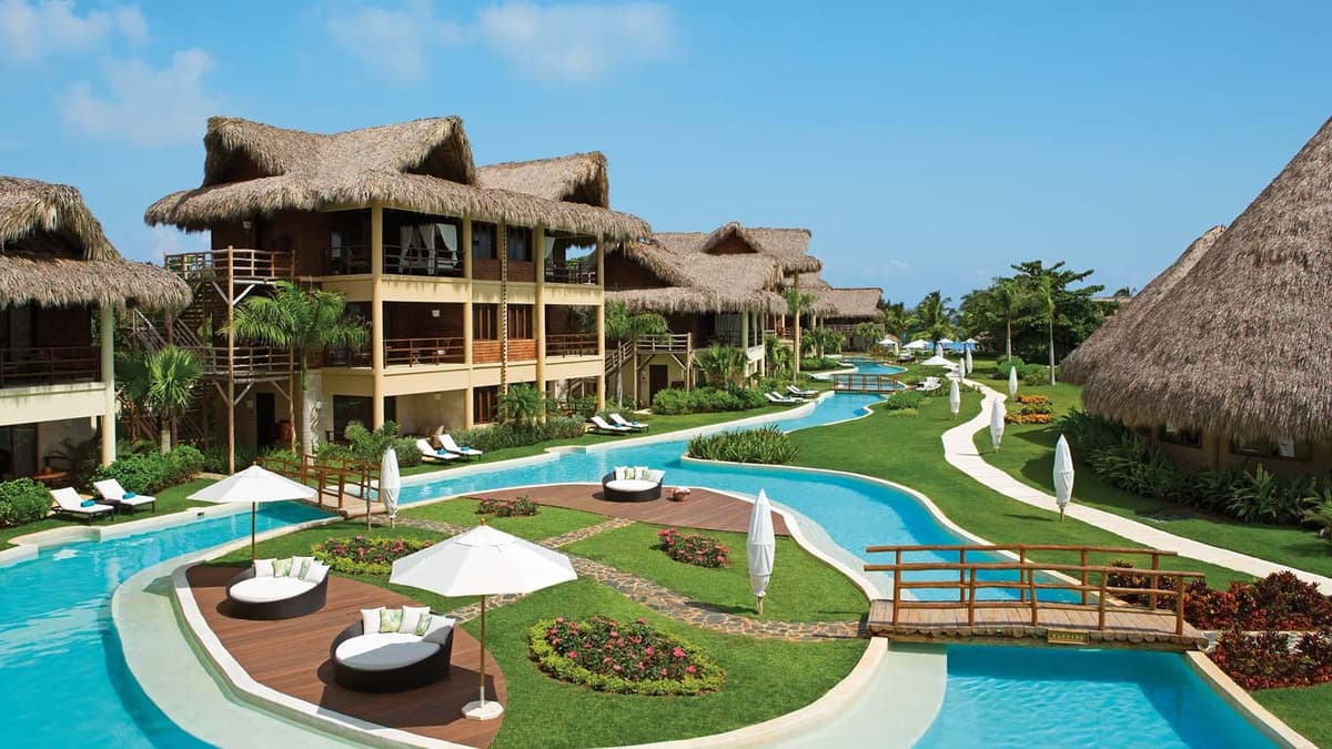 Serpentine pool with Balinese daybeds and umbrella-shaded loungers winding past palapa-roofed suites