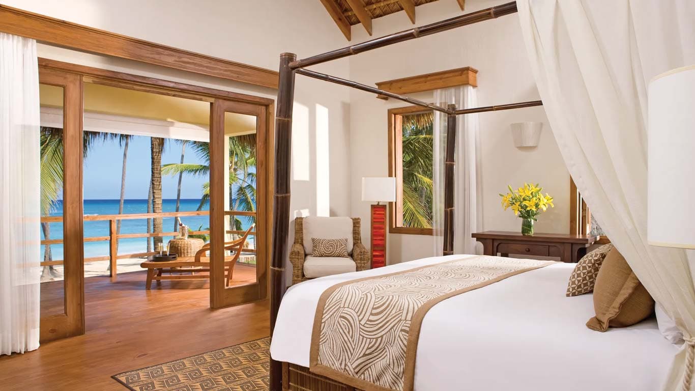 Villa suite bedroom with four-poster canopy bed, panoramic ocean views through French doors, and palapa ceiling